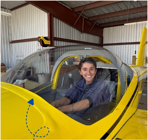 Leilani smiling in the cockpit of a small yellow airplane inside a hangar