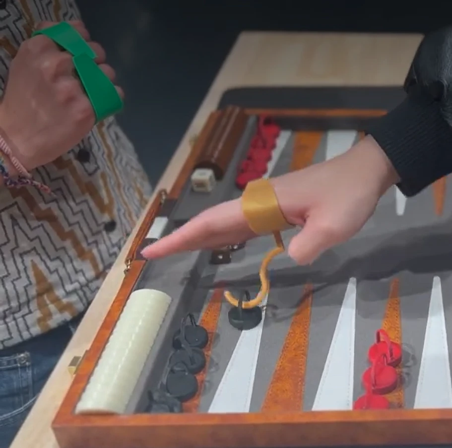 A hand wearing a gold finger ring with a hook that lifts a game piece by its loop handle on a wooden game board, while another player wears a green hand strap