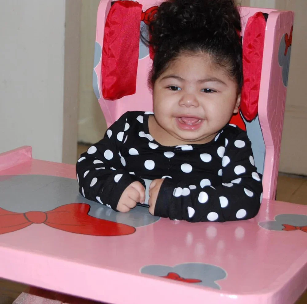 A smiling young child sitting in a pink custom-built adaptive chair with a tray and red side supports