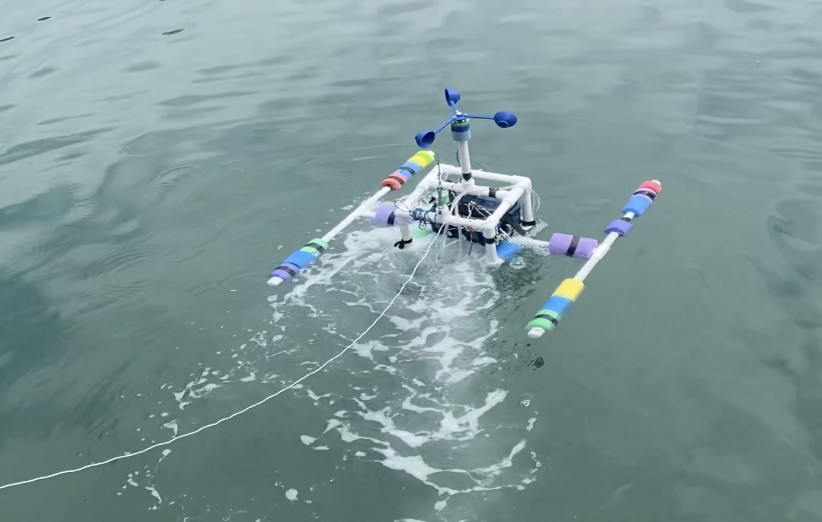 The surface robot in the ocean: a PVC frame with pool-noodle outrigger floats, a waterproof electronics box, and an anemometer on a mast, leaving a wake
