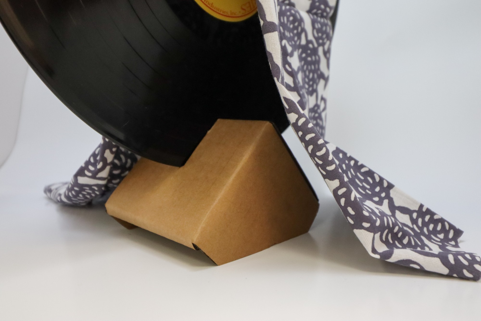 A folded cardboard packaging corner reused as a stand holding a vinyl record, draped with a patterned tenugui cloth