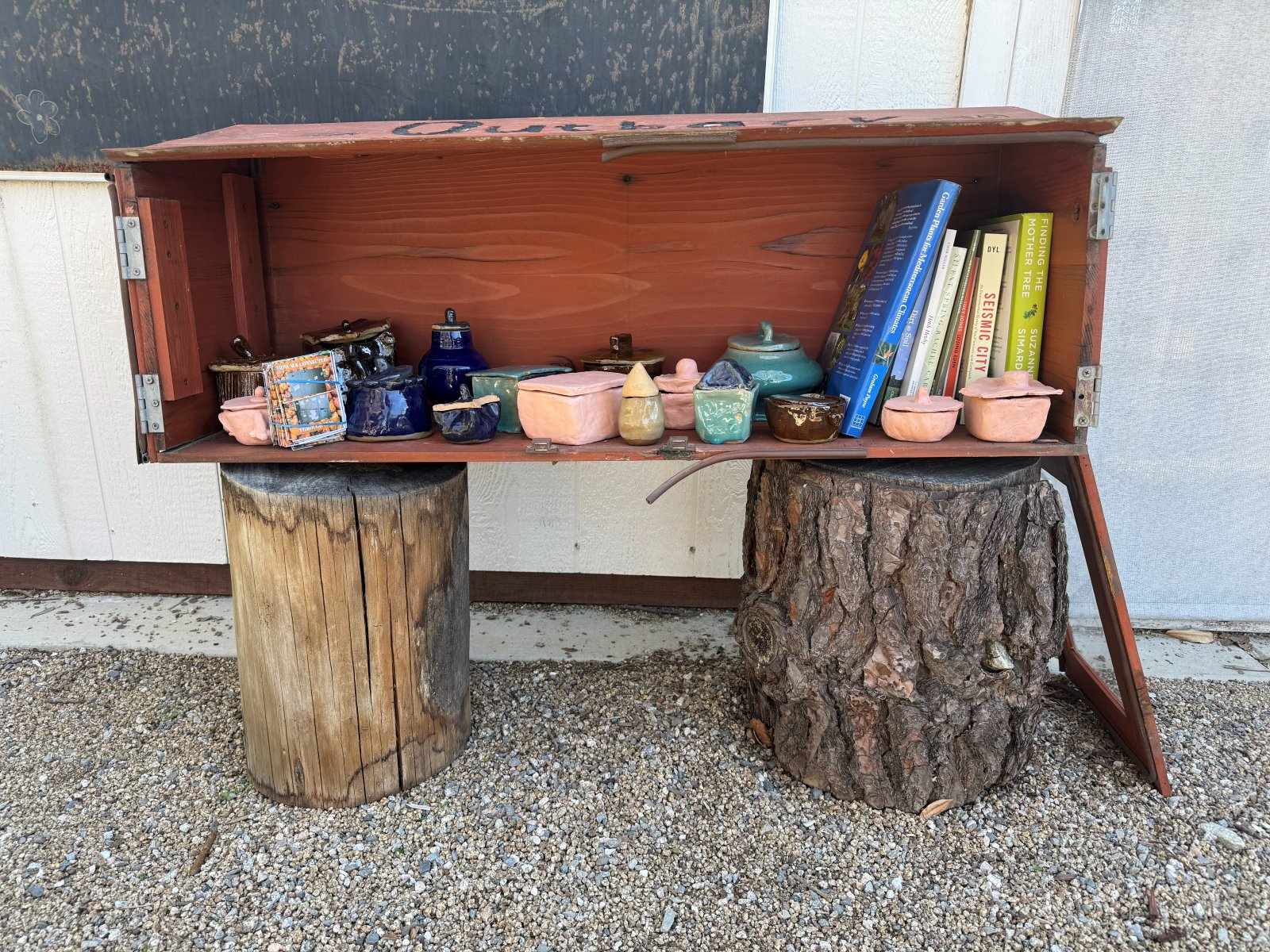 Handmade ceramic lidded pots lined up in a red wooden shelf box beside garden books, set on two tree stumps