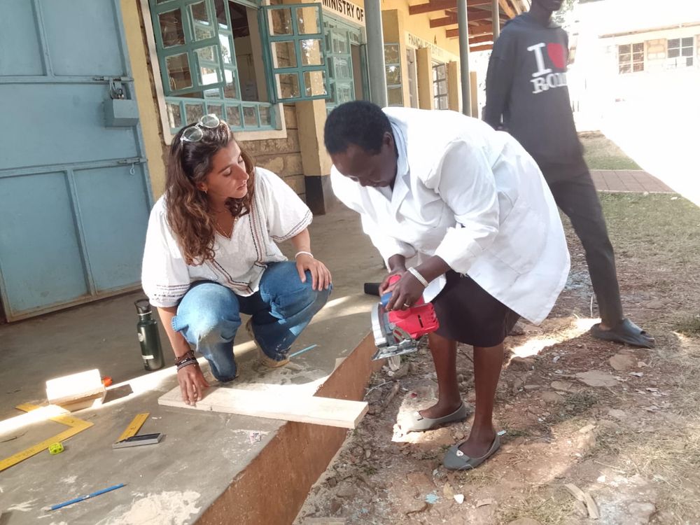 Leilani kneeling beside a teacher using a circular saw to cut wood on a workbench outside the school
