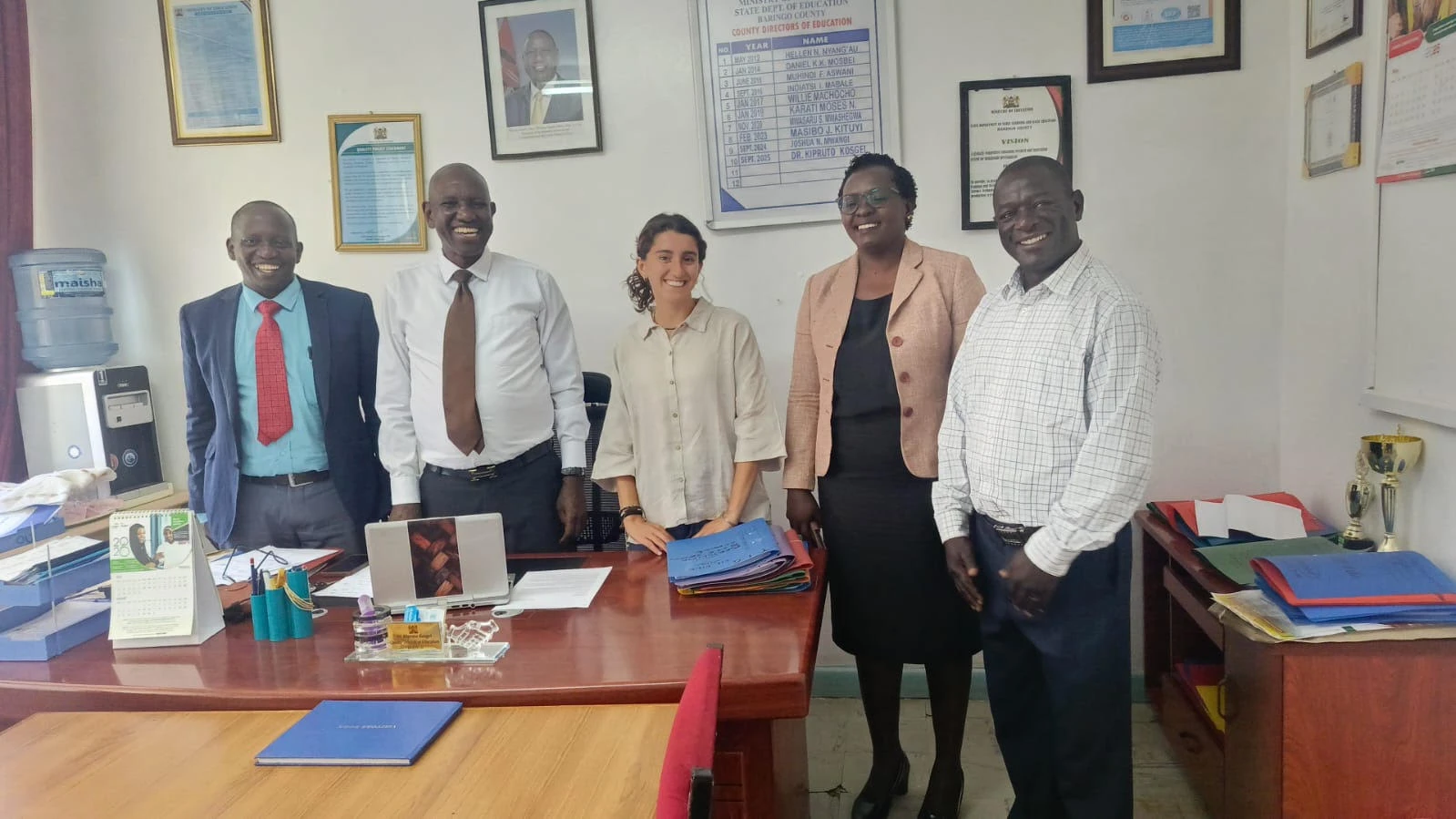 Leilani smiling with Dr. Kosgei, Baringo County Minister of Education, and three other officials around a desk in the Baringo County Ministry of Education office