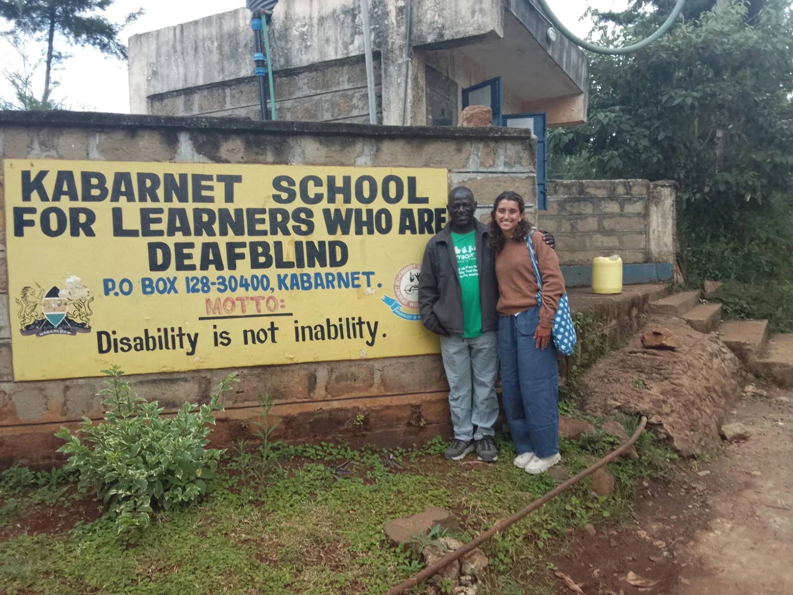Leilani and teacher Jared Rakiro standing beside the painted sign for Kabarnet School for Learners Who Are Deafblind, whose motto reads 'Disability is not inability.'