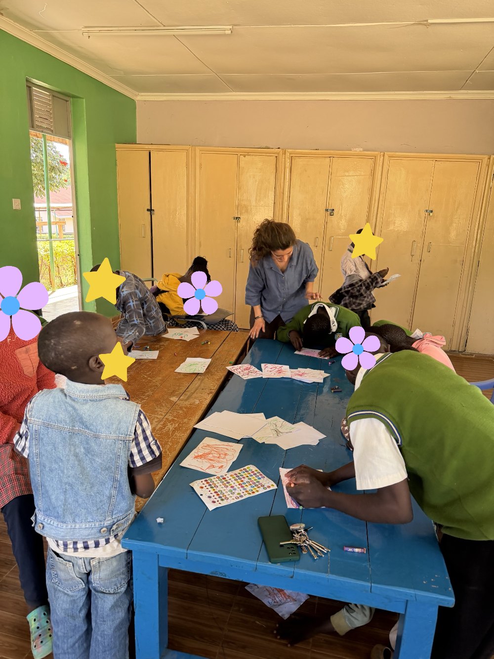 Learners drawing with crayons at two long tables during art class while Leilani leans in to help; learners' faces are covered with flowers and stars for privacy