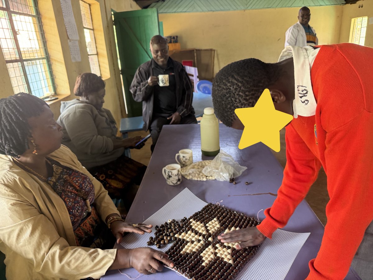 Teachers and a learner around a purple table, running their hands over a beaded mat of dark and light wooden beads strung onto plastic canvas; the learner's face is covered with a star for privacy