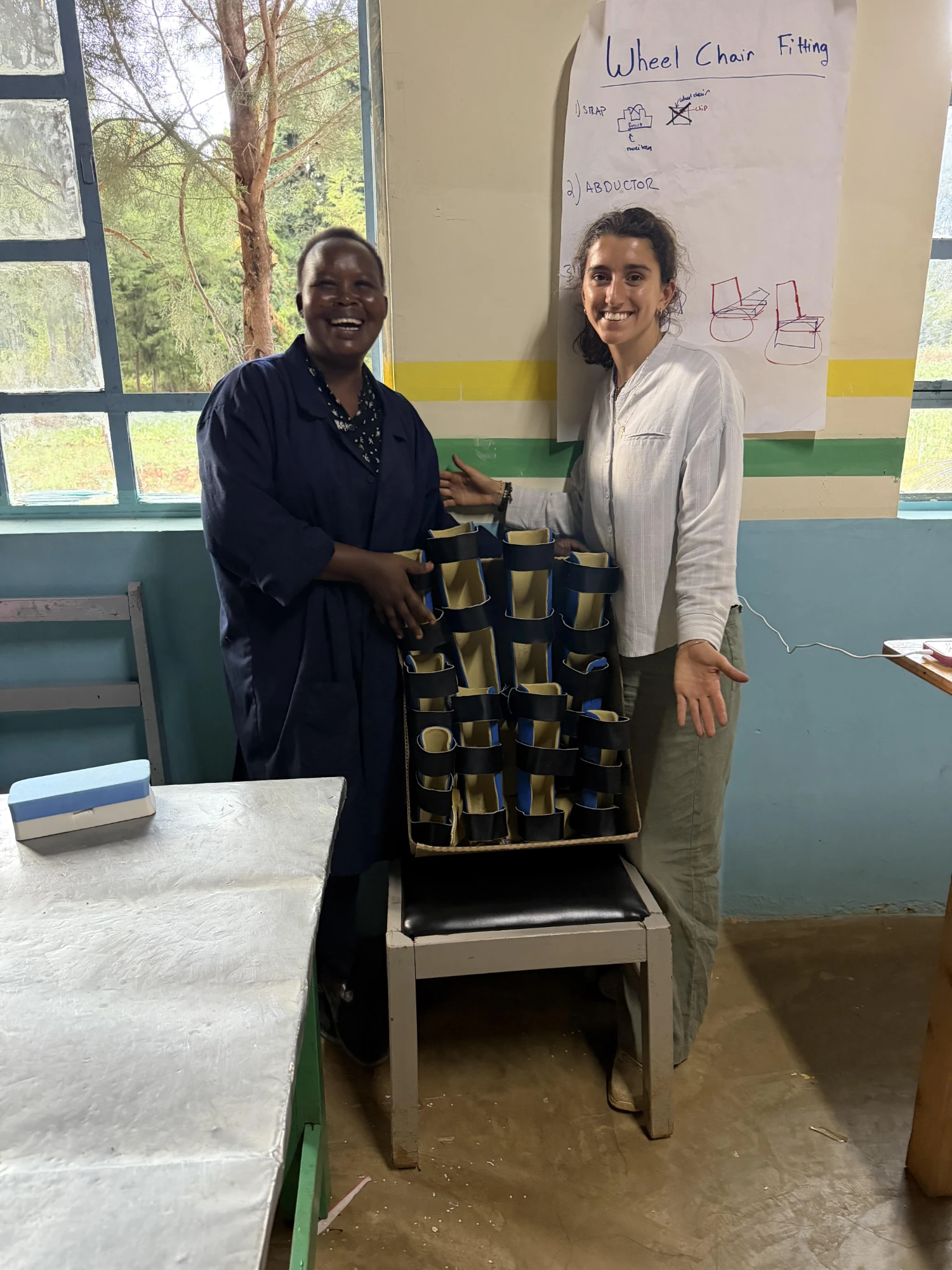 Leilani and Eddah smiling beside a box full of finished leg and arm braces with blue padding and black straps, in front of a hand-drawn wheelchair fitting chart