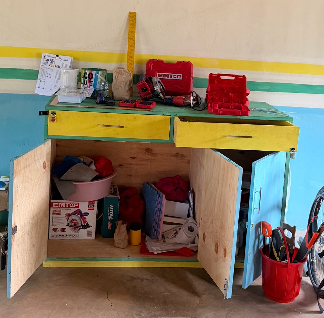 The tool cabinet Leilani built, painted green, yellow and blue, with its doors and a drawer open to show power tools, supplies and a bucket of hand tools beside it