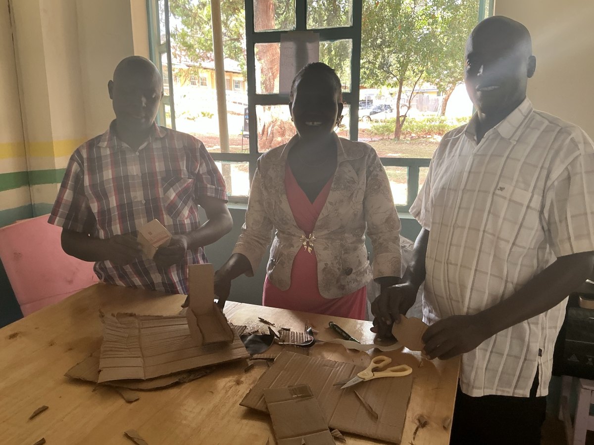 Three teachers at a workbench covered in cut cardboard, holding up the cardboard mockups they made of their project ideas