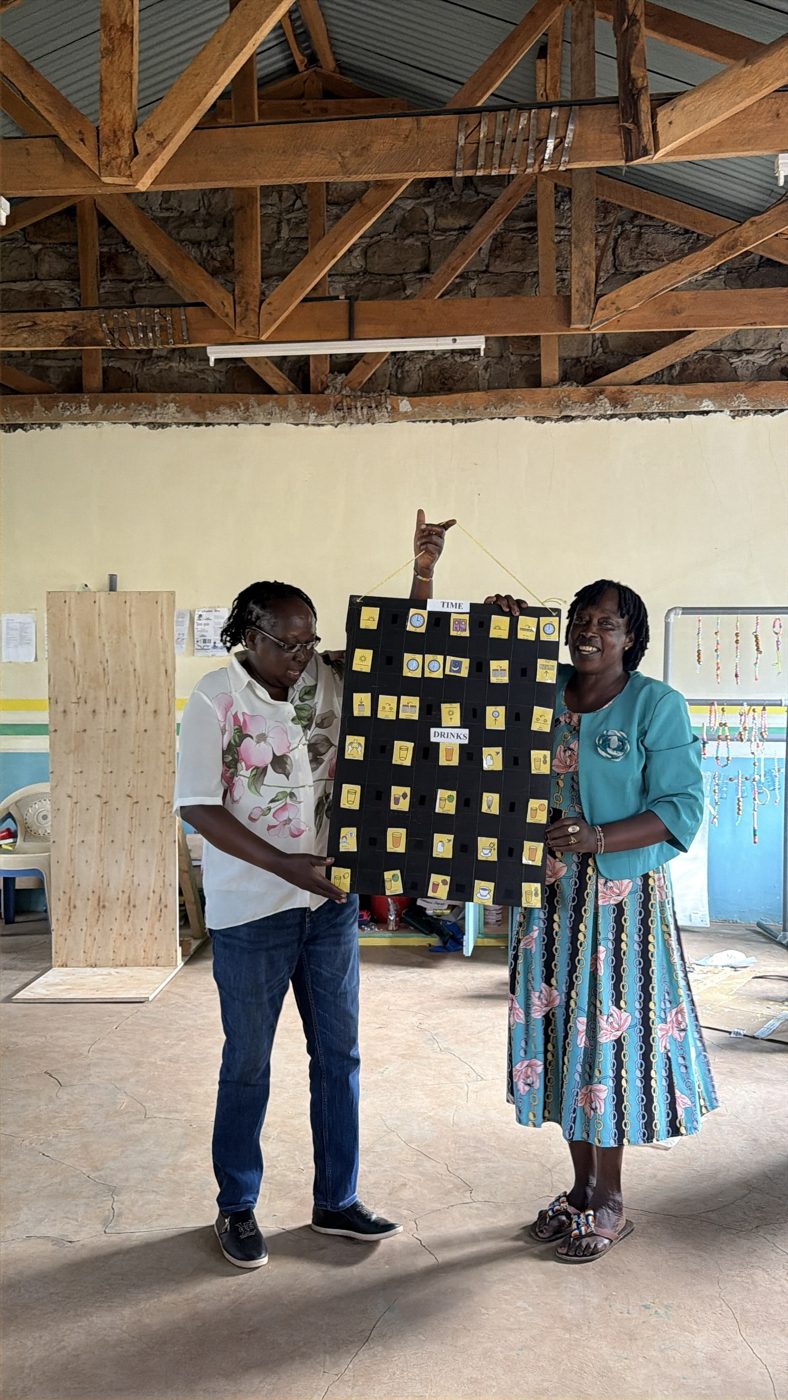 Two teachers holding up a hanging communication board: a black grid of yellow picture symbol cards sorted under Time and Drinks headings