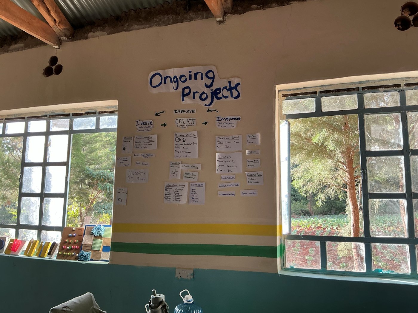 The Ongoing Projects wall in the workshop: a design cycle of Ideate, Create, Implement and Improve, with paper cards for each project, such as the rocking chair, texture board, numbers board and wheelchair fitting, placed under the stage it has reached