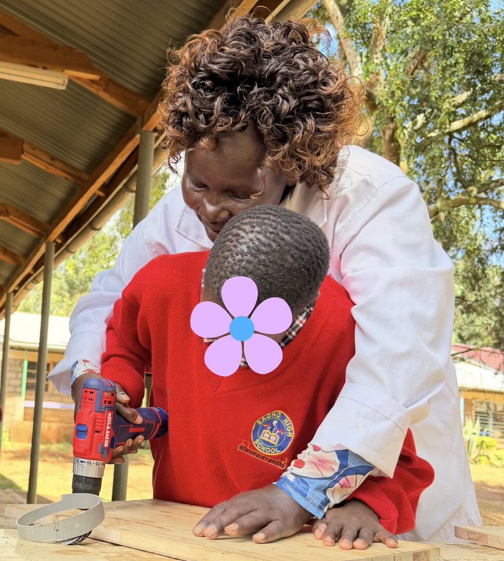 A teacher in a white coat standing behind a learner in a red sweater, guiding her hands as she drills into a wooden board; the learner's face is covered with a flower for privacy