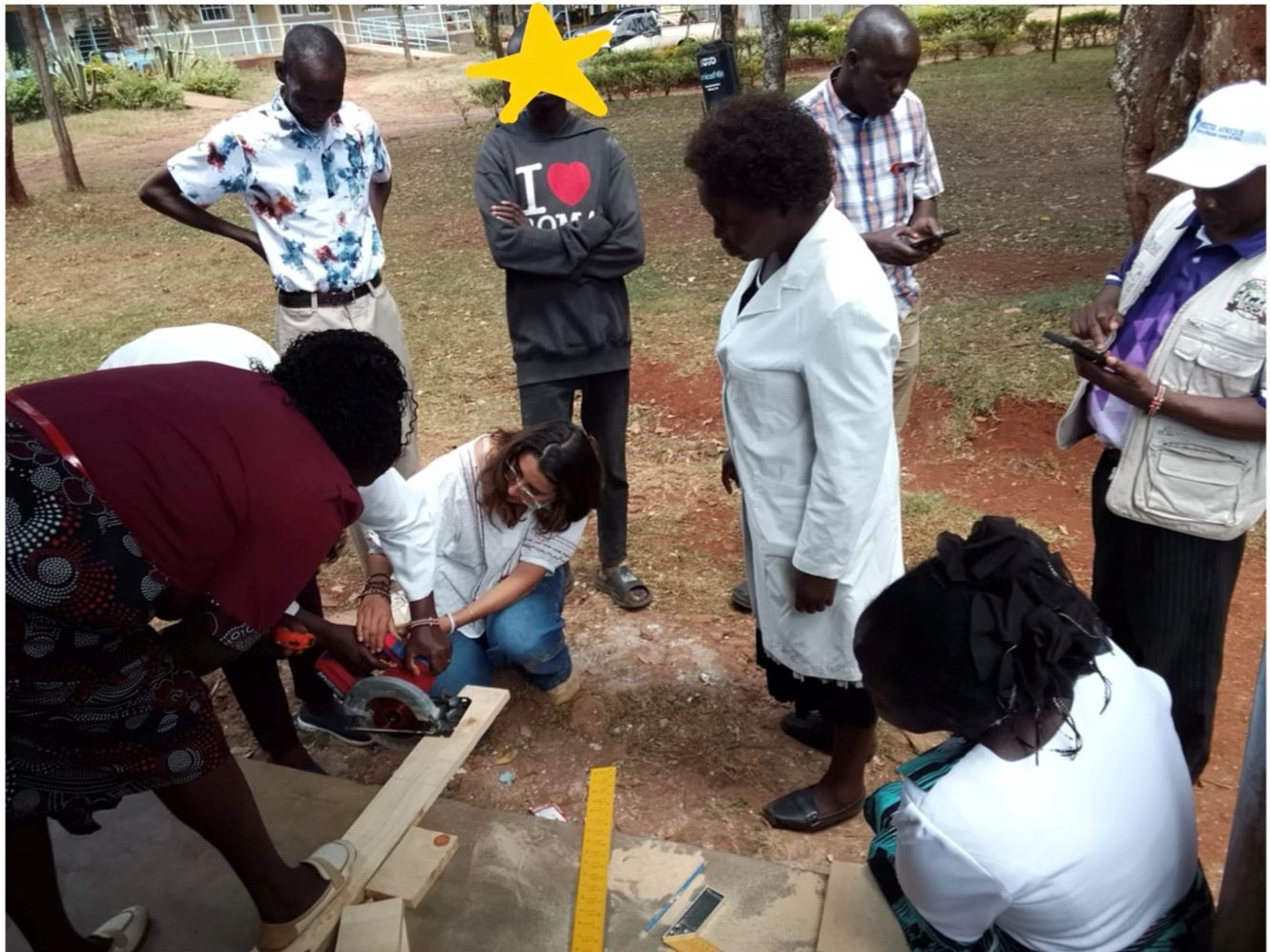 A woman teacher using a circular saw on a wooden board outdoors while Leilani kneels beside her, guiding and steadying the saw, as other teachers and staff look on; one learner's face is covered with a star