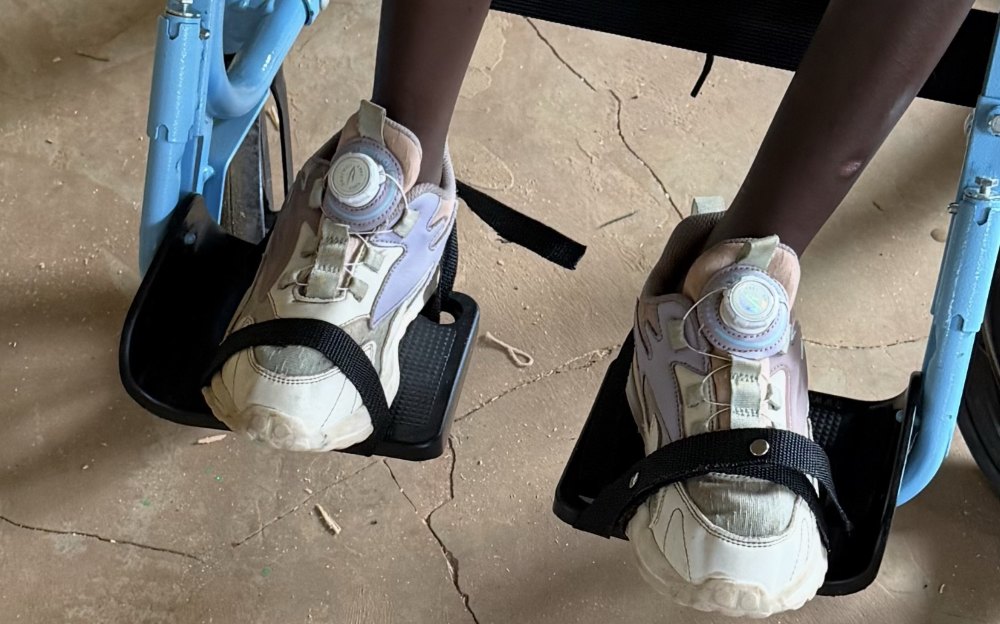 A learner's sneakers held in place on a wheelchair's footrests by black webbing foot straps