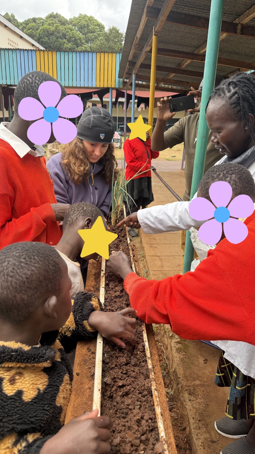 Learners, Leilani and a teacher gathered around a raised garden bed made from a reclaimed rain gutter, pressing seeds into the soil beside a row of green onions; learners' faces are covered with flowers and stars for privacy