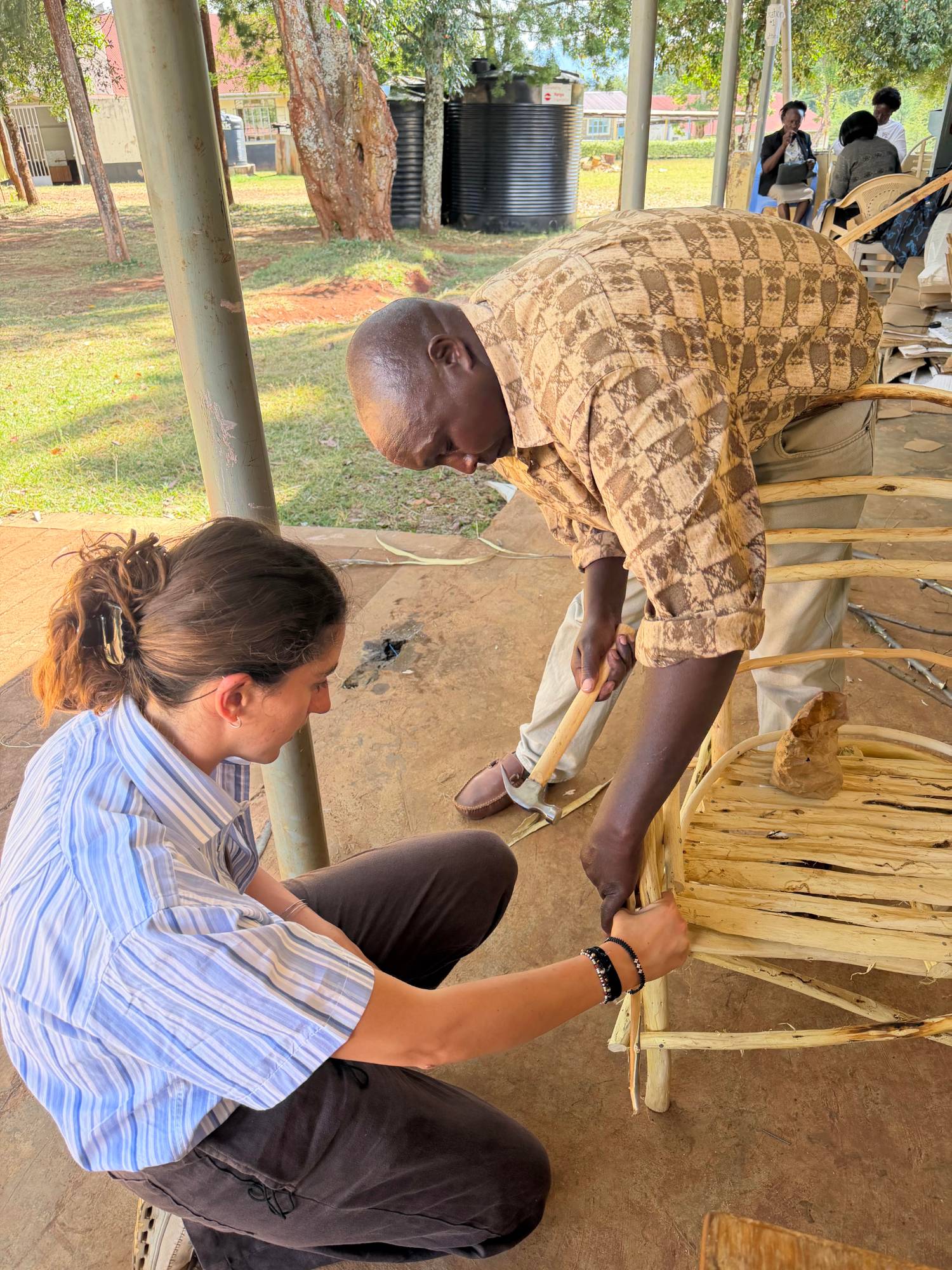 Leilani crouching to hold a joint steady while a local maker hammers together a chair made from peeled cypress branches