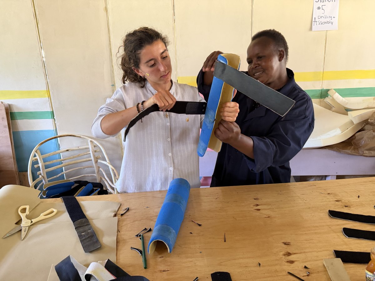 Leilani and a woman in navy work clothes holding up a blue-padded PVC backslab to fit its black hook-and-loop strap, with a finished brace and foam scraps on the workbench