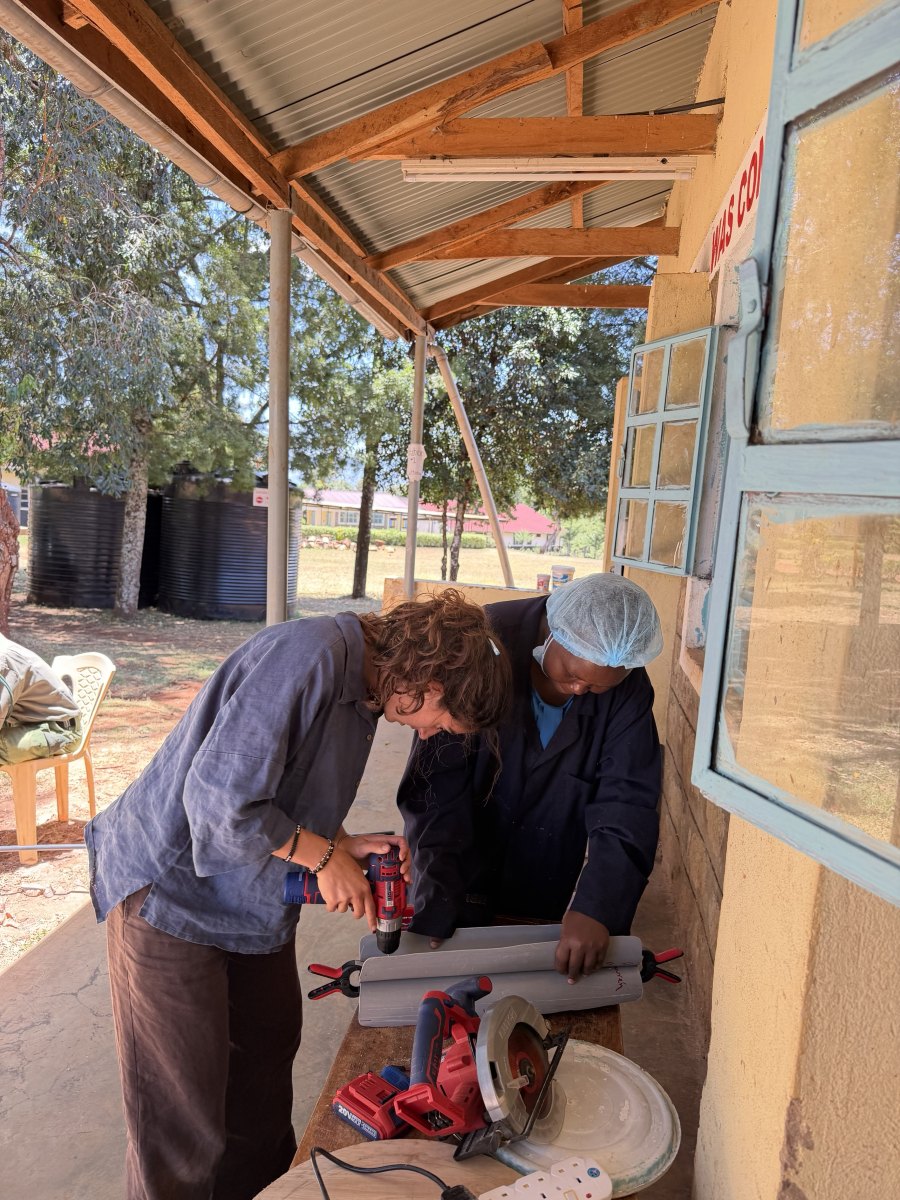 Leilani drilling into a length of gray PVC pipe clamped to a bench outdoors while a woman in a hairnet and navy work coat holds it steady, with a circular saw on the bench