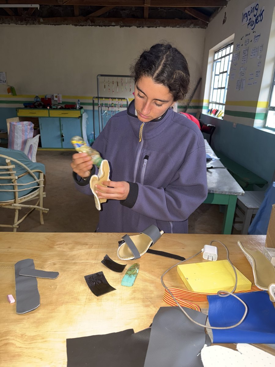 Leilani gluing a padded lining into a white Orthoplast foot splint at a workshop table covered in straps, vinyl and hook-and-loop scraps, with a finished strapped splint in front of her