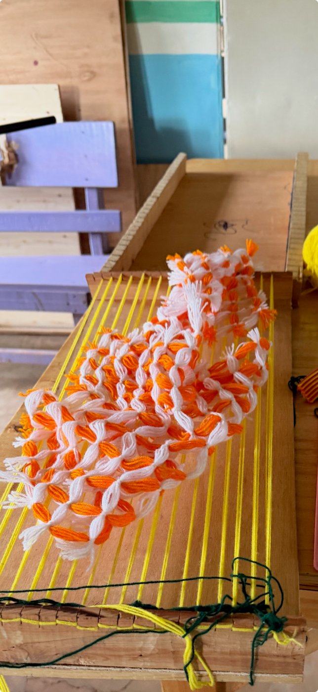A wooden frame loom strung with yellow warp threads, holding a piece of orange and white yarn weaving