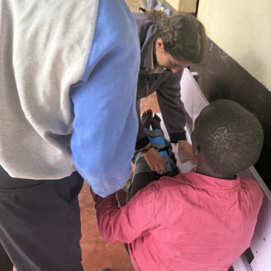 Leilani fastening the blue-padded mermaid brace around a learner's legs as the learner lies on a bench, with a teacher and another learner looking on