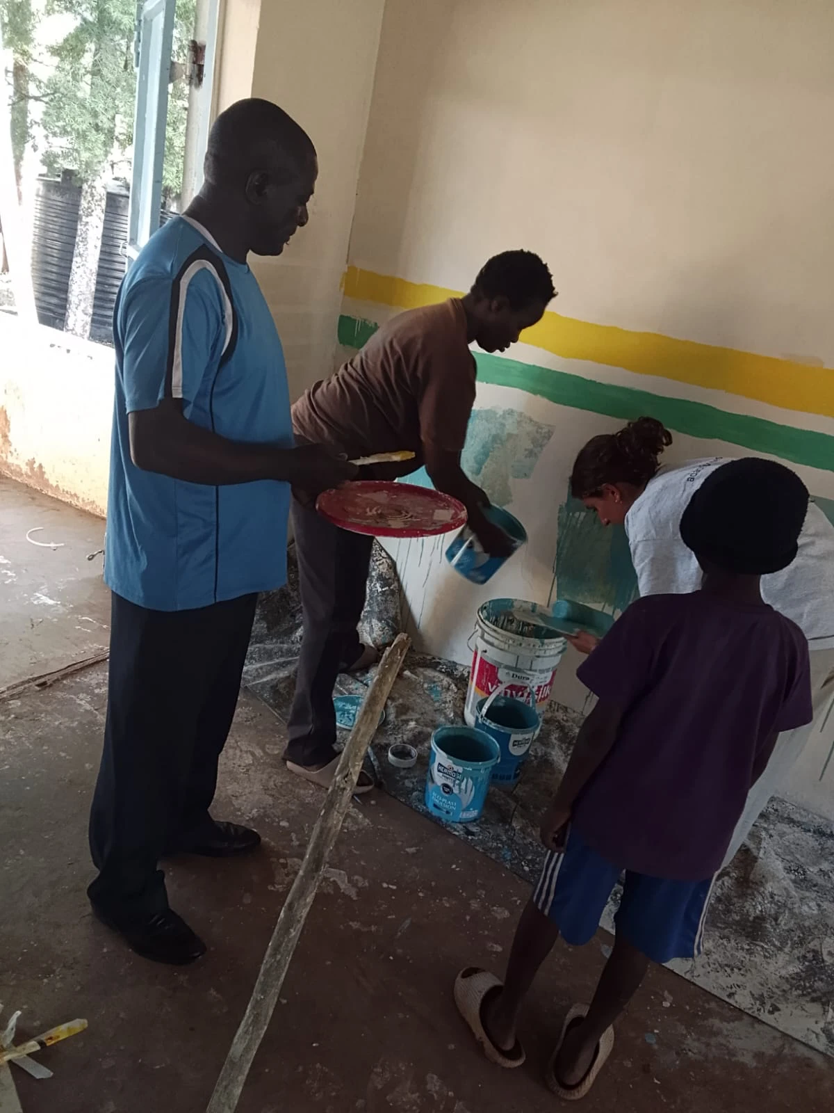 Leilani painting a teal wall inside the empty workshop room, with a man and a boy helping and paint buckets on the floor