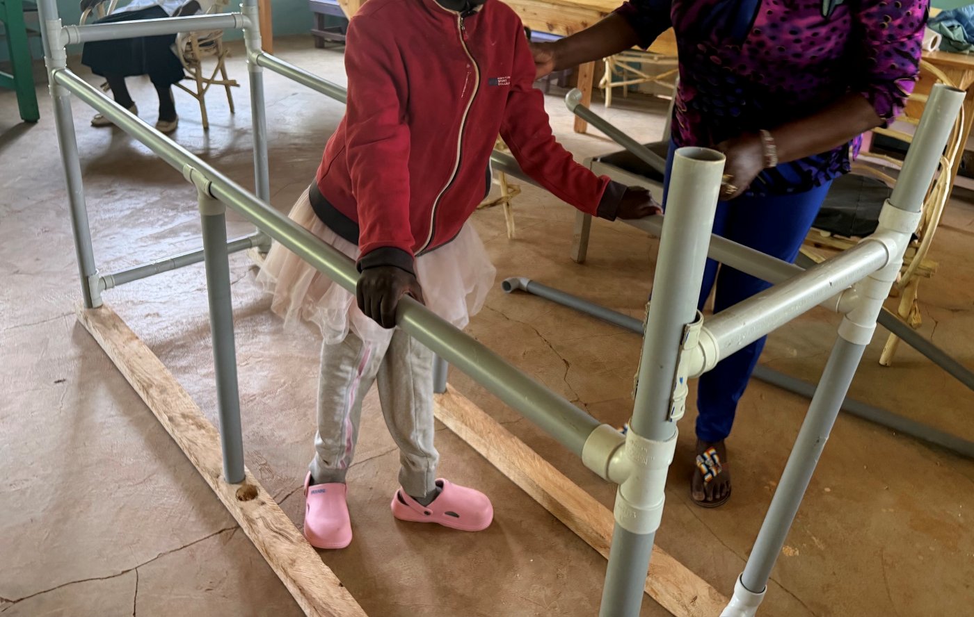 A learner in pink shoes walking between PVC-pipe parallel bars on a wooden base, holding the rails while a teacher steadies her