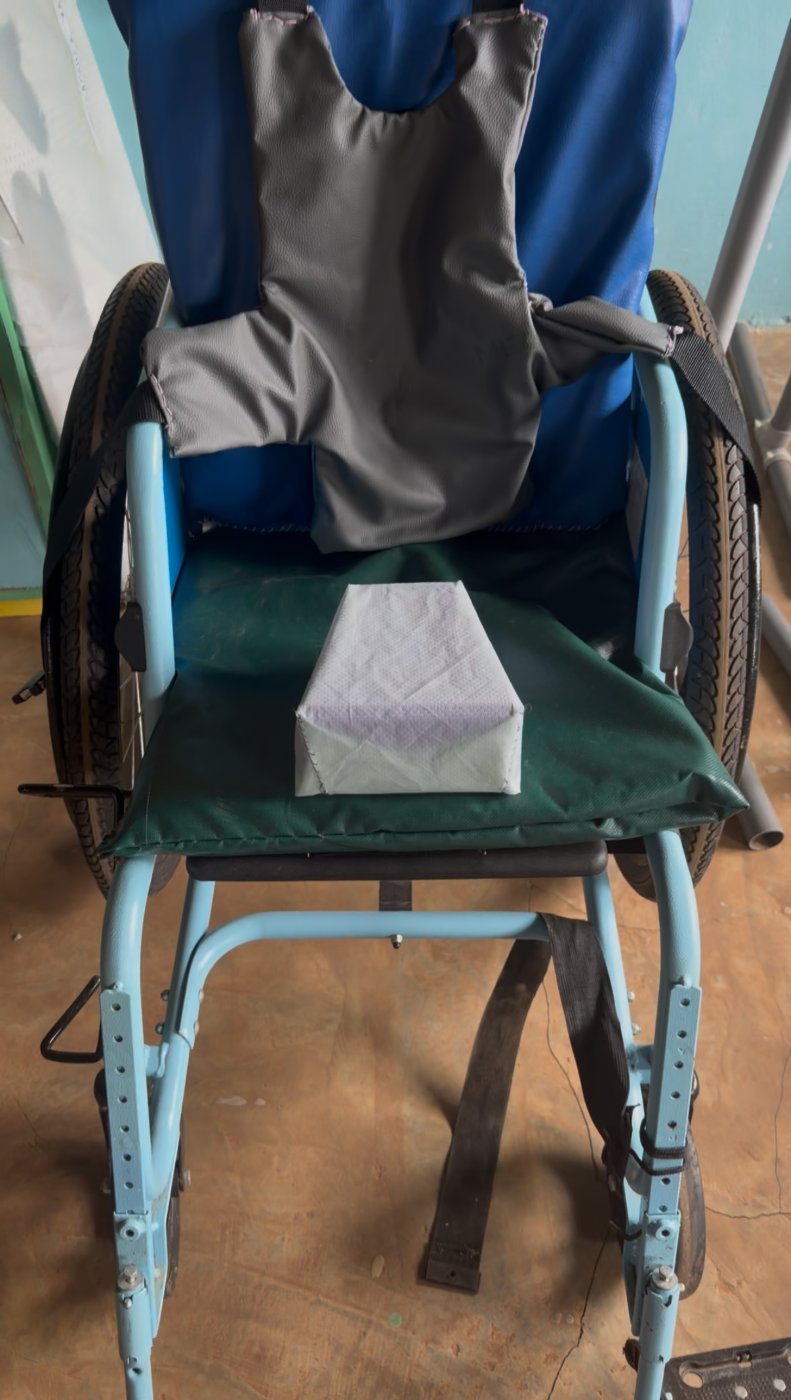 A wheelchair fitted with a padded gray chest harness and a fabric-covered pommel (abductor) cushion on the seat