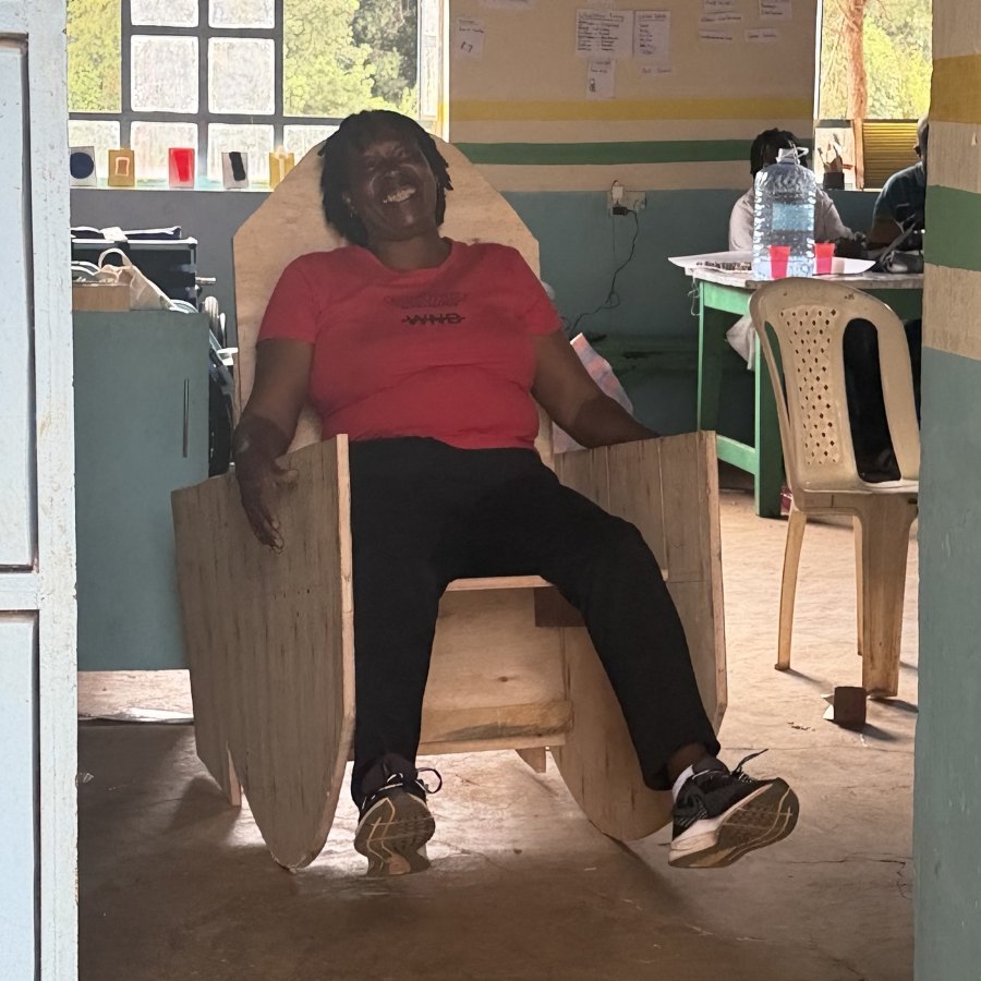 A teacher laughing as she leans back in the finished plywood rocking chair in the workshop, under the Ongoing Projects wall
