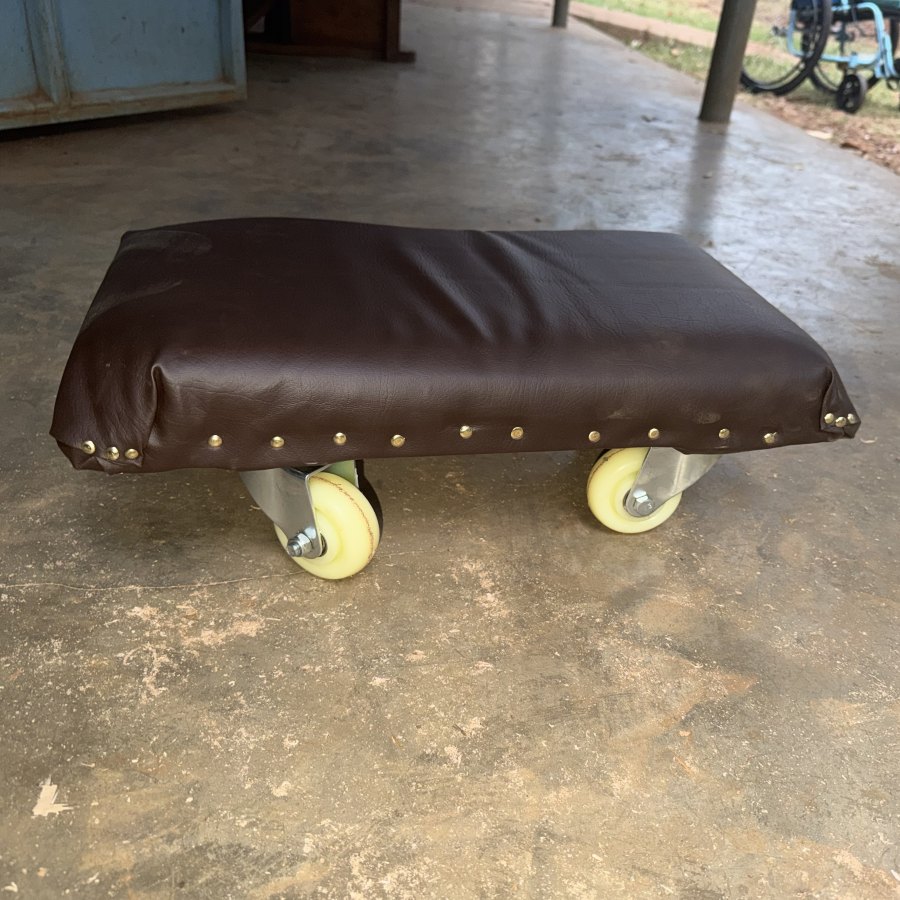 A low scooter board: a padded brown vinyl seat finished with brass tacks, on four caster wheels