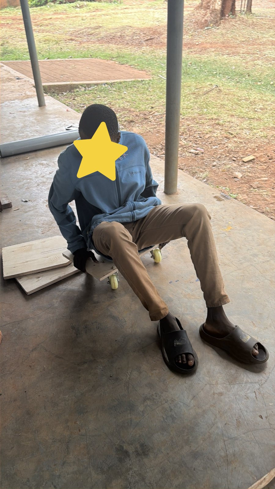 A learner sitting on a low wooden scooter board with caster wheels, pushing along the concrete floor with his feet; his face is covered with a star for privacy