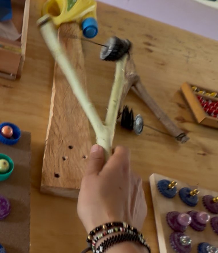 A hand holding a forked branch shaker strung with bottle caps that rattle when shaken, on a workshop table beside counting boards