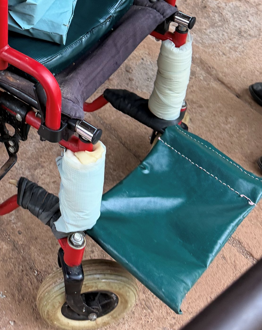 A wheelchair fitted with a soft green vinyl sling footrest, with its footrest posts padded and taped