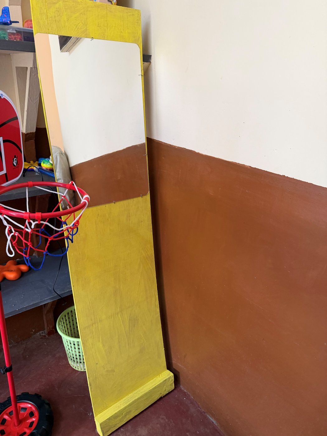 A tall speech mirror: a mirror panel set into a yellow-painted wooden board standing on a base, leaning against the classroom wall