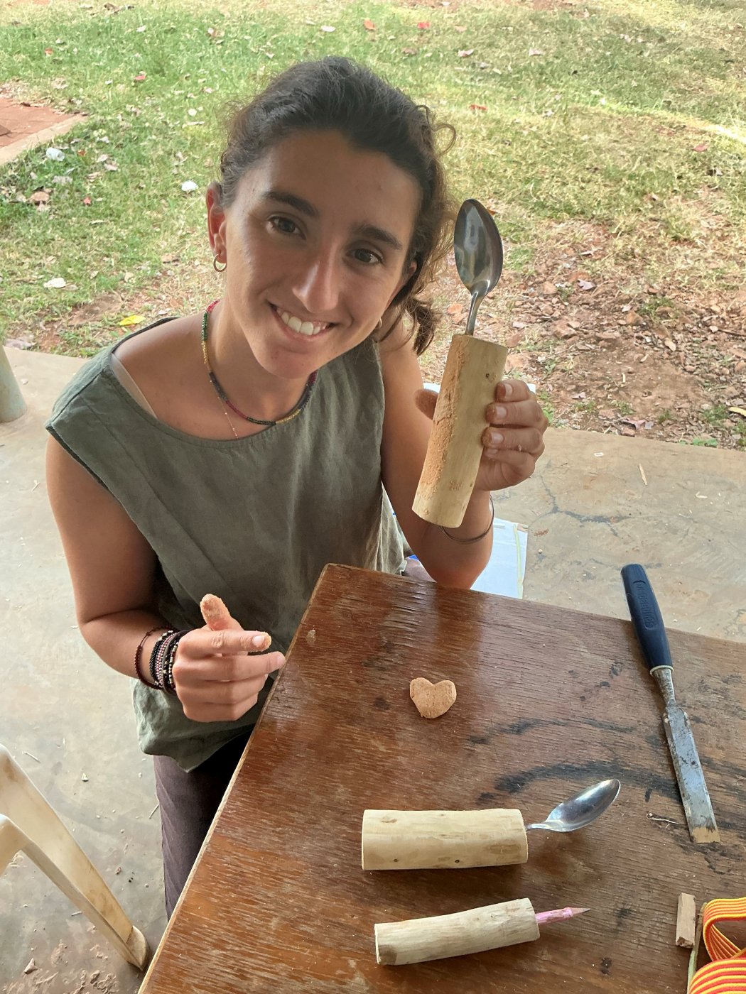 Leilani smiling and holding up a spoon set into a thick wooden branch handle, with more built-up spoon and pencil handles and a chisel on the table