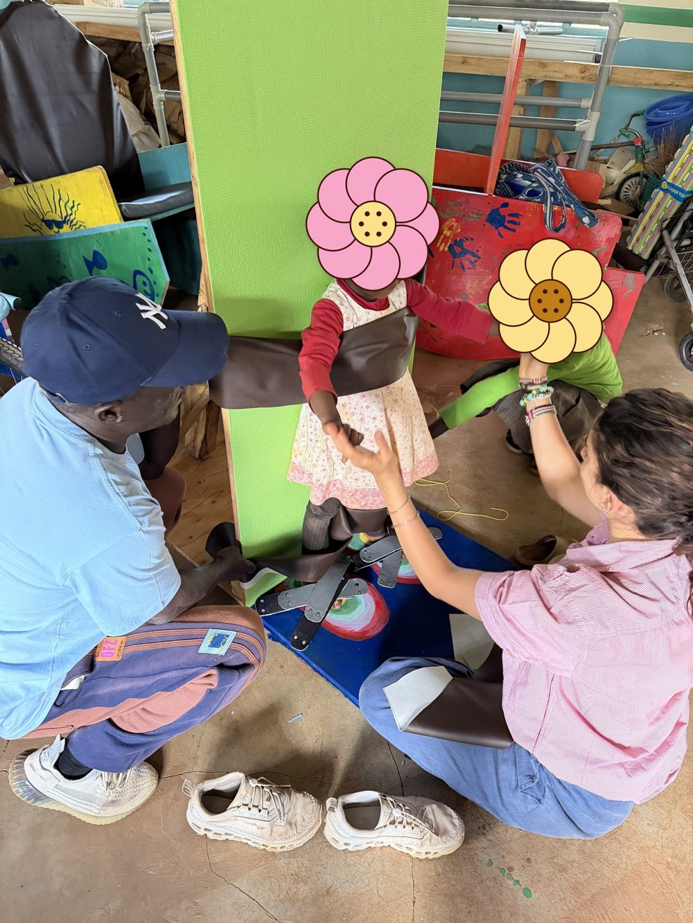 A learner standing strapped to the green padded standing board, with chest and knee straps and hinged foot straps on a painted blue footplate, holding hands with Leilani while a teacher adjusts a strap; learners' faces are covered with flowers for privacy