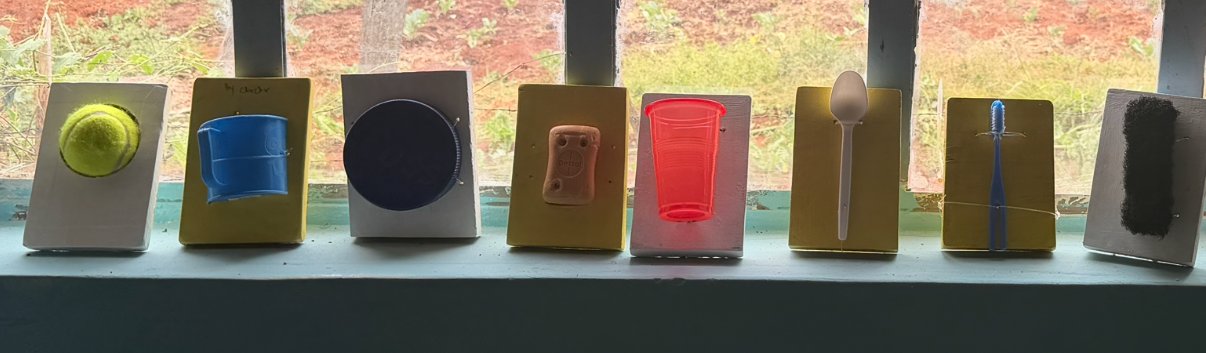 Eight tactile cue cards on a windowsill: a tennis ball, cup, jar lid, bar of soap, plastic cup, spoon, toothbrush and scrub sponge, each mounted on a painted wooden block