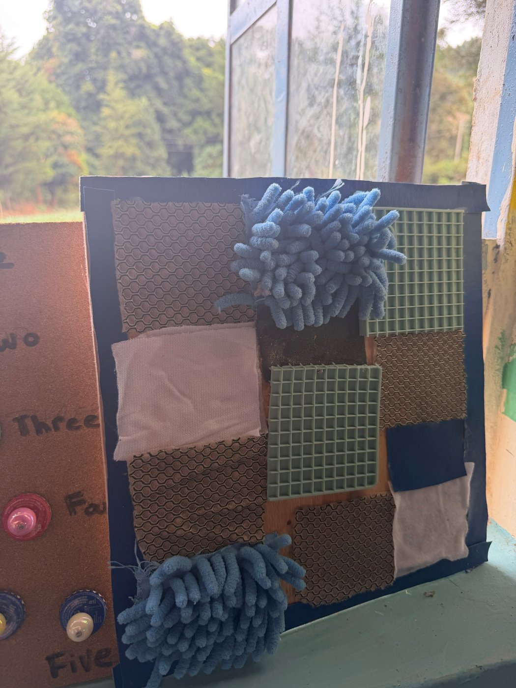 A sensory texture board covered in patches of different materials: shaggy blue microfiber, rubber grid mats, ridged mesh and soft white fabric