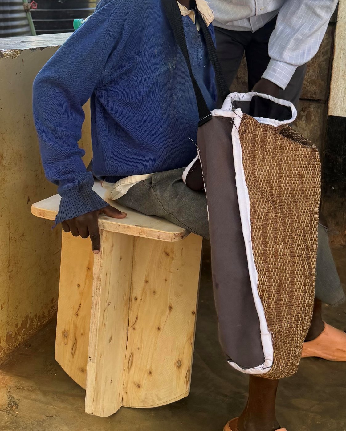 A learner sitting on a simple wooden tippy stool, with a teacher standing beside them