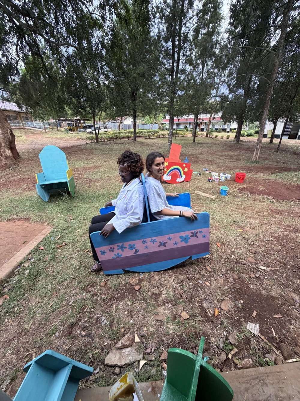 Leilani and a teacher sitting back to back, smiling, in a two-person plywood rocker painted blue and mauve with flowers and butterflies, outside under the trees