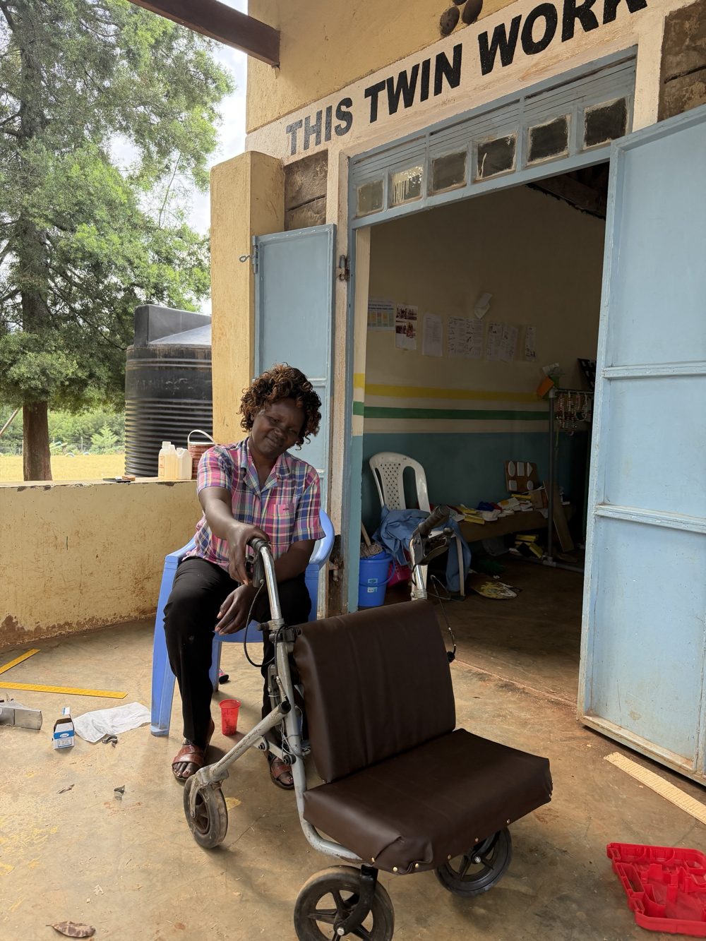A teacher seated beside a rolling walker converted into a scooter, with a padded brown vinyl seat and backrest fixed to the frame