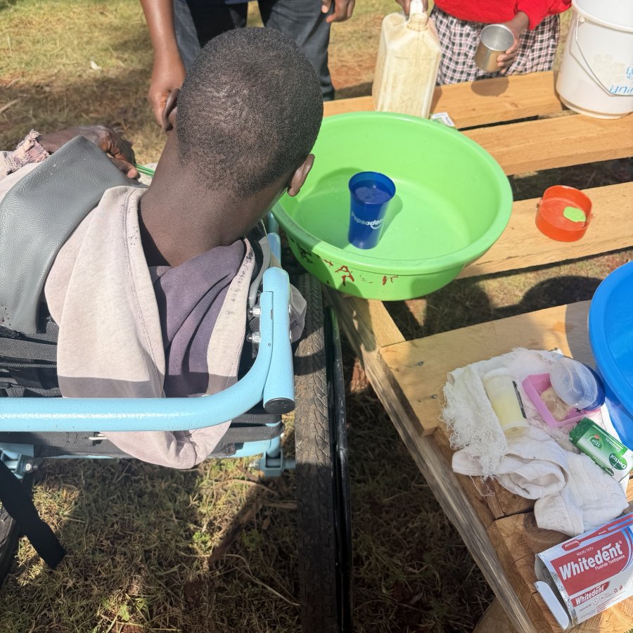 A learner in a wheelchair, seen from behind, pulled up to a wooden washing table holding a green basin, a cup, soap and toothpaste, while a teacher and another learner help