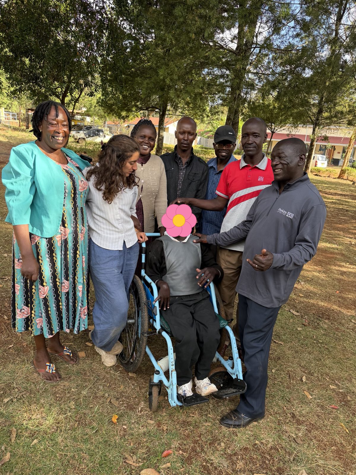 Leilani and a group of adults standing under the trees around a smiling learner in a light blue wheelchair who is wearing a gray padded chest harness, with hands resting on his head and shoulder; his face is covered with a flower for privacy