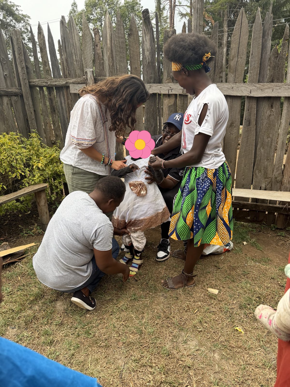 Leilani, a teacher and family members gathered around the laughing girl in her white party dress, holding her hands and steadying her as she stands in her leg braces in the family's yard; her face is covered with a flower for privacy