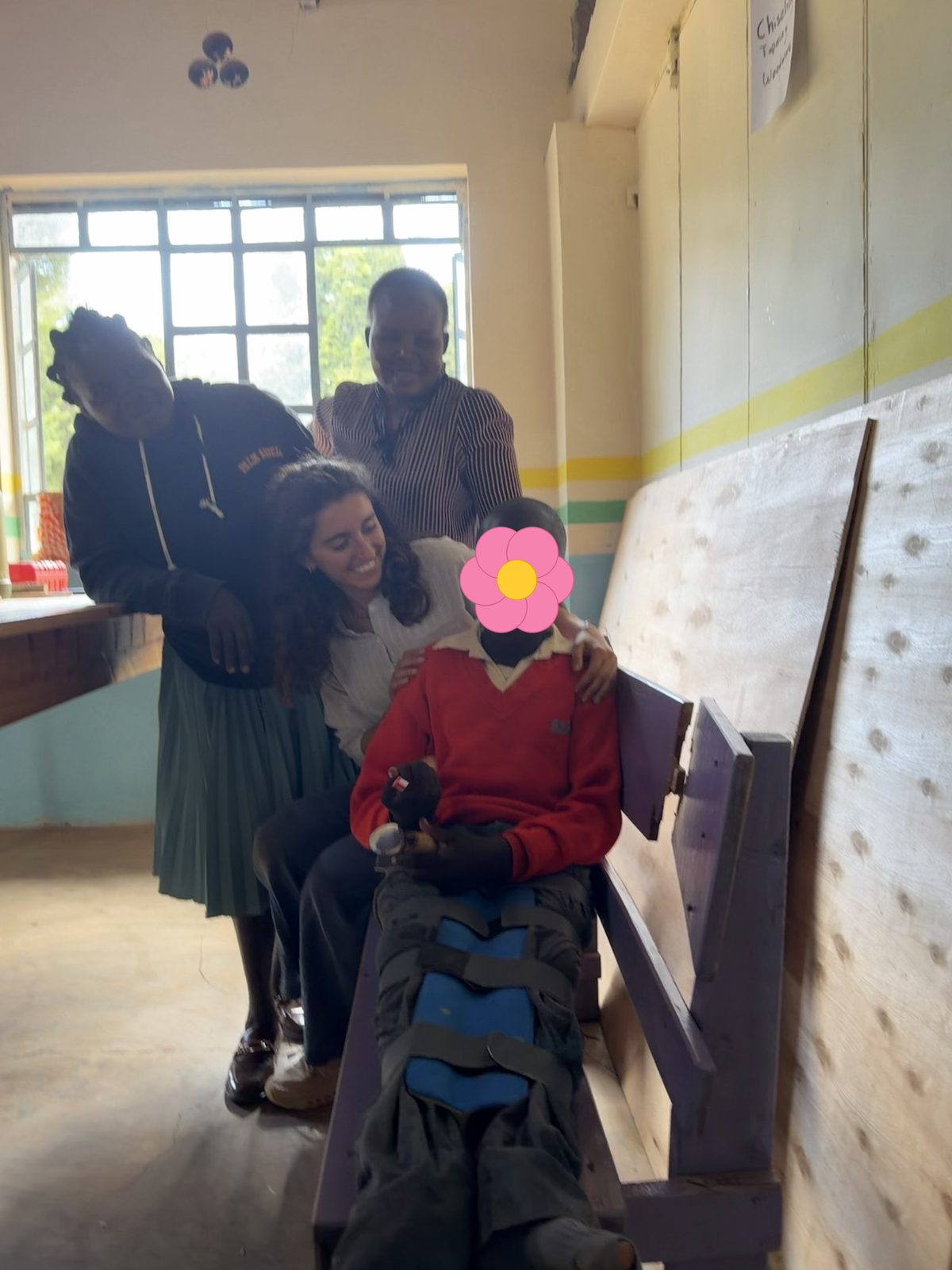 Leilani hugging a smiling learner in a red school sweater who sits on a purple bench wearing a blue padded knee brace and holding his built-up spoon and toothbrush, with his mother and another woman leaning in behind them; his face is covered with a flower for privacy