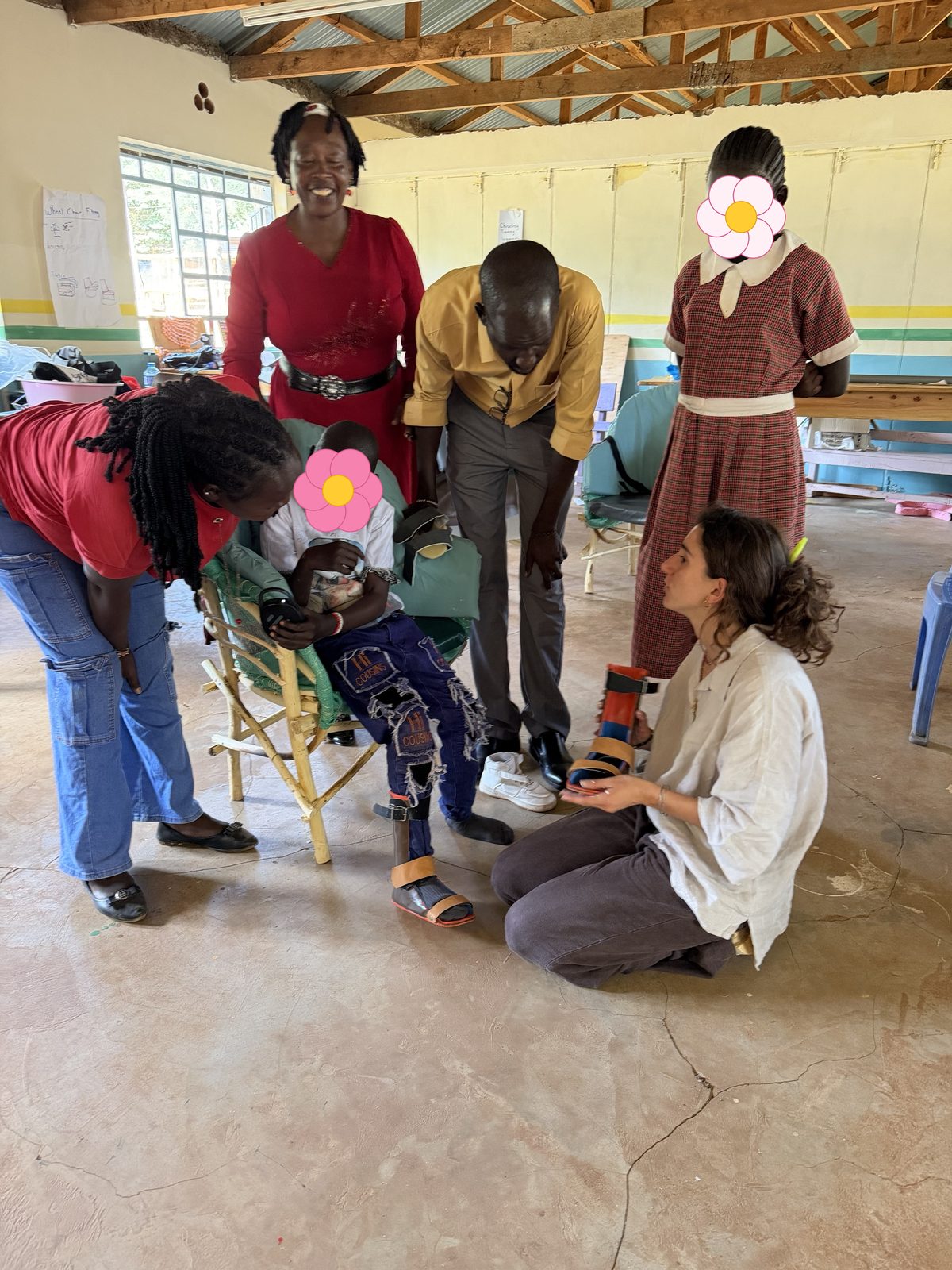 Leilani kneeling on the workshop floor holding up an orange and blue leg brace, explaining it to a learner wearing a matching brace in a cypress-branch chair while adults and another learner look on; the learners' faces are covered with flowers for privacy