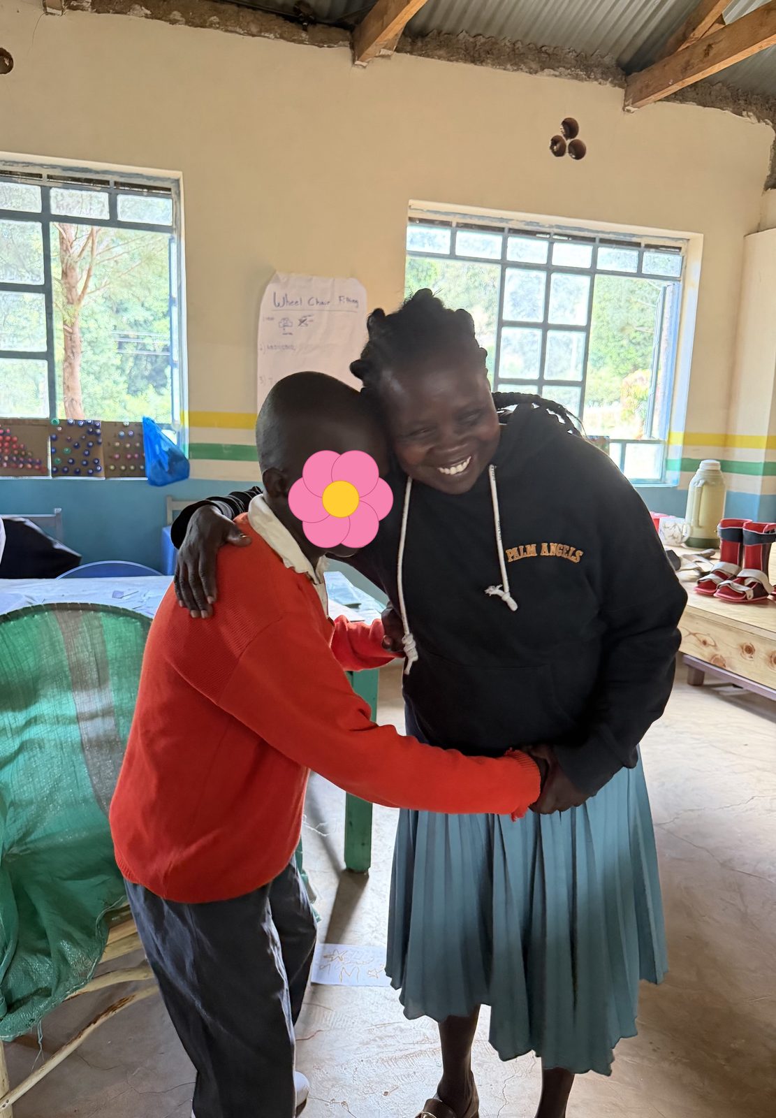 A learner in a red school sweater standing and hugging his smiling mother in the workshop after being fitted with his new braces; his face is covered with a flower for privacy