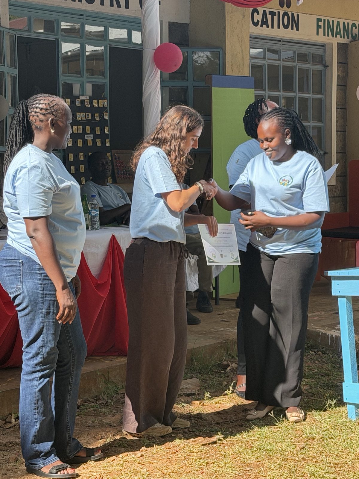 Leilani shaking hands with a smiling teacher as she presents her adaptive design certificate outside the school, with another teacher beside them and the communication board hanging behind