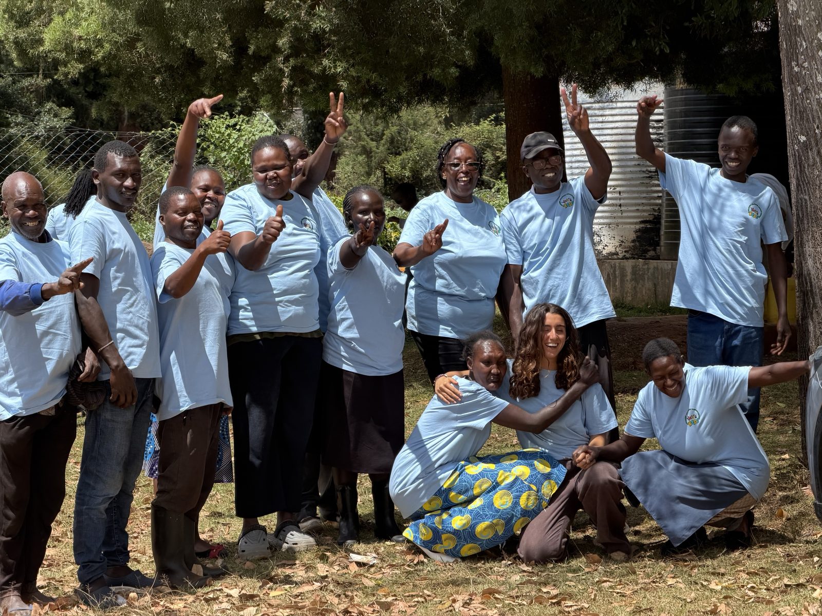 The graduating teachers in matching light blue workshop T-shirts cheering, pointing and giving thumbs up under the trees, with Leilani crouching in front hugging two teachers